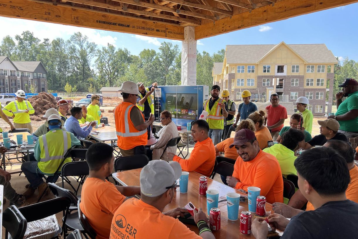Construction workers eat lunch together at picnic tables underneath a wooden awning 