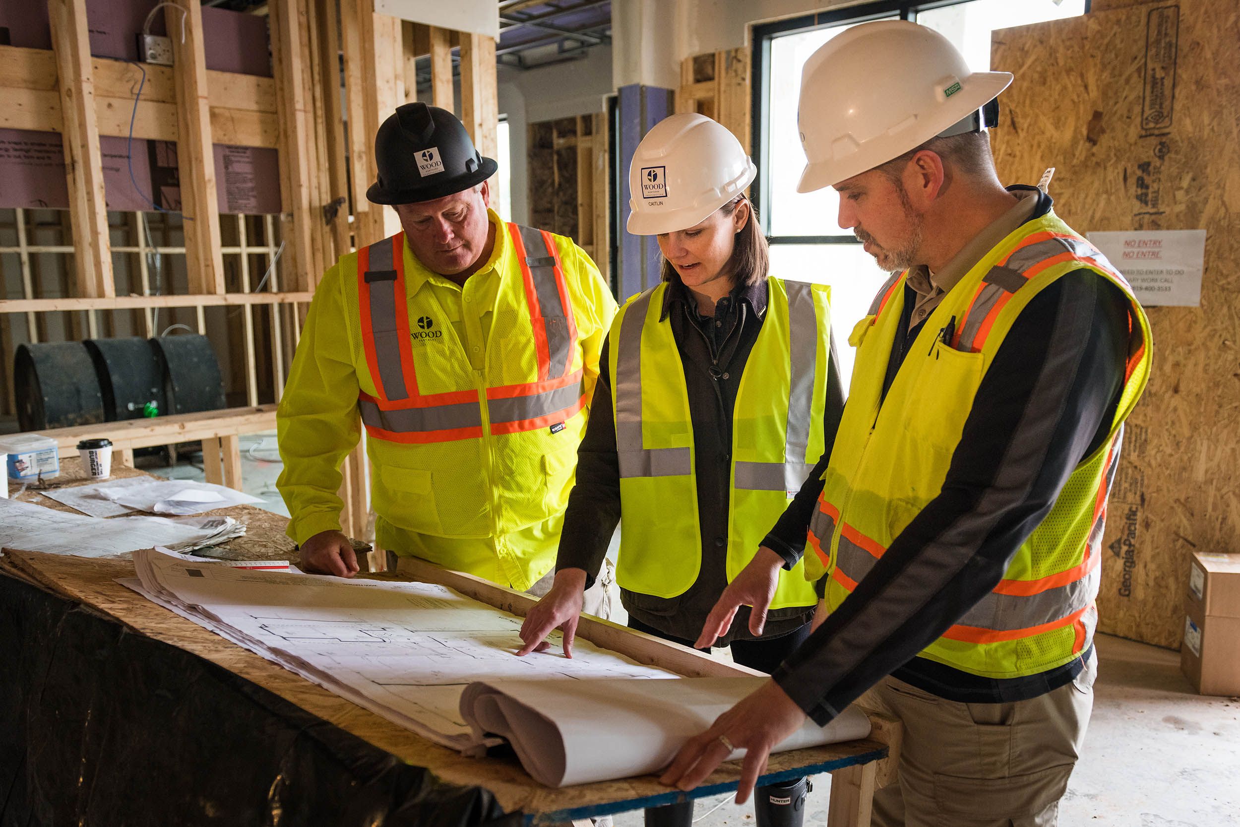 Two men and one woman in hard hats and construction vests reviewing blueprints